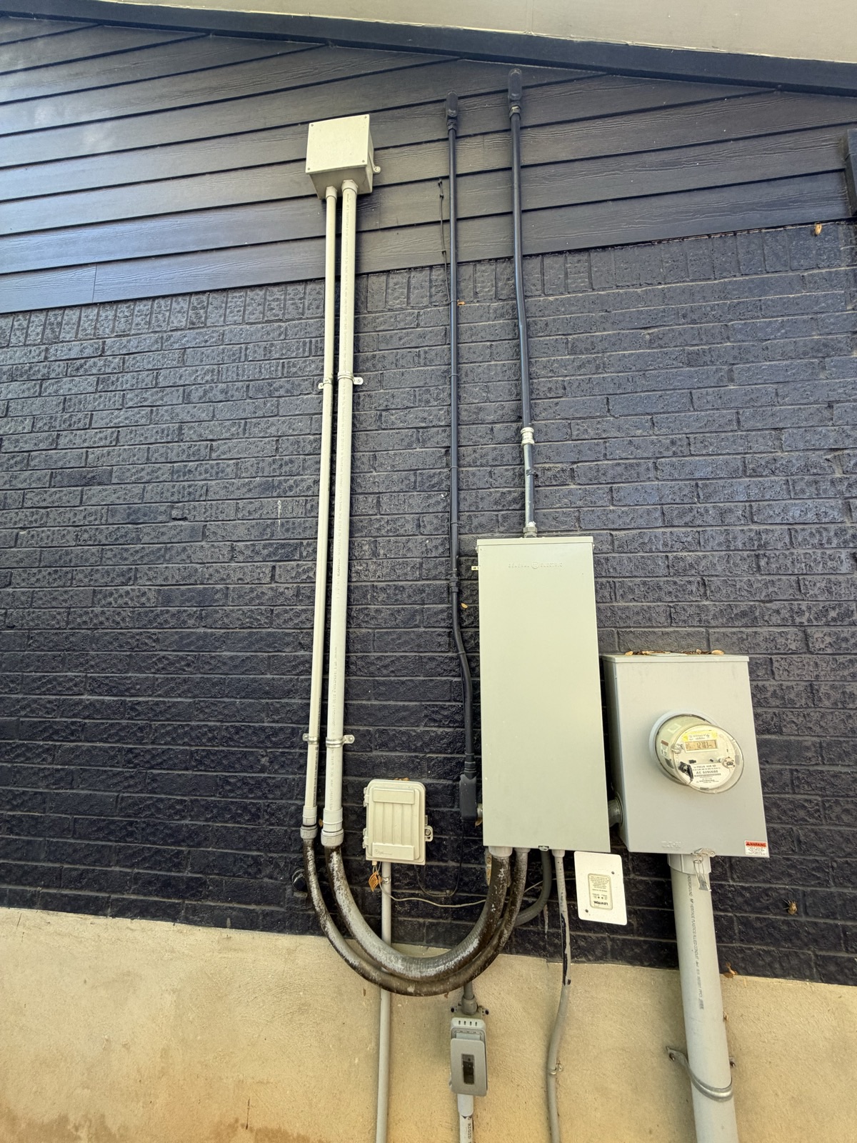 Photo of the exterior brick wall with the main panel and meter, with the new conduit drawn in: a 3/4 in. PVC run from a top knockout on the panel straight up the brick and siding to an LB fitting that turns into the attic, held by two-hole straps.